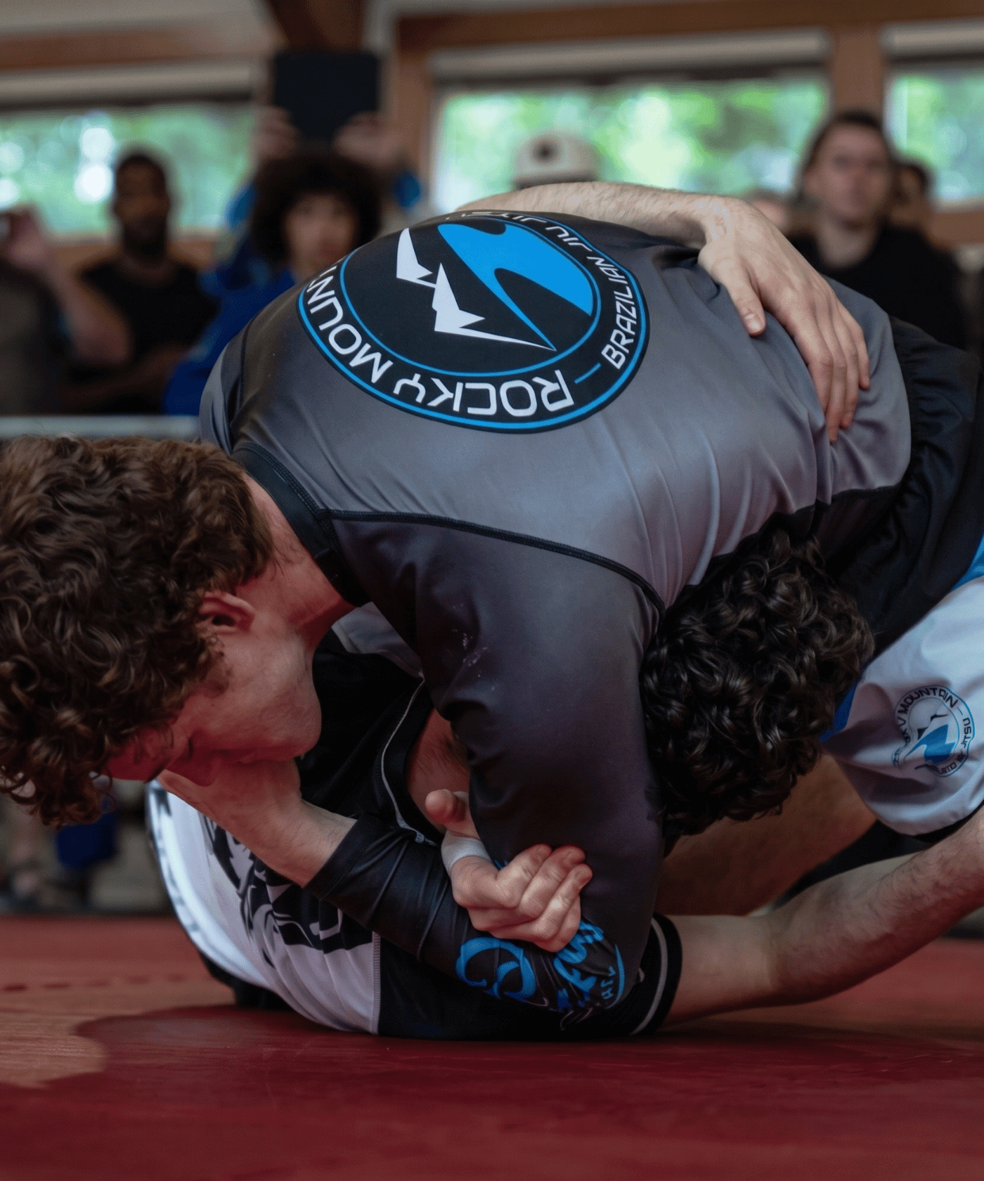 Two individuals in a grappling position on a red mat with 'Rocky Mount' branding.